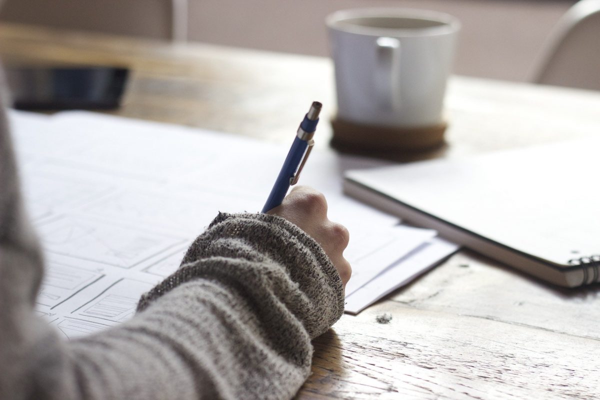 A person writes notes on a sheet of paper; a coffee cup and notebook are in the background.