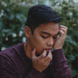 A side profile of Miggy, who crouches amidst bushes and white flowers that recede blurrily into the background. His fingers gently crawl up from his long-sleeved maroon shirt, over his chin and lips, and toward his black, wavy hair. The brown skin of his cheek is caressed by his palm as he looks down with eyes closed in contemplation.