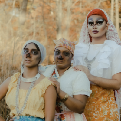 Three women in front of a field of beige crops. The women on the left and the middle are on the same height and have a black makeup around their eyes and noes, while the woman on the right has a fairer makeup and is in a taller stance than the other two.